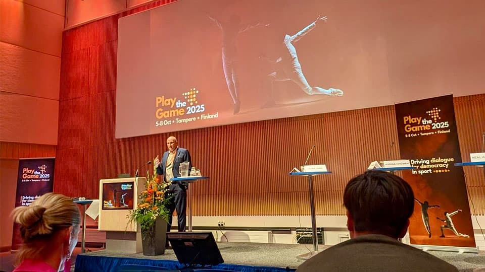 Alumnus Neal standing on stage with a screen in the background that reads Play the Game 2025. 5-8 October. Tampere. Finland.